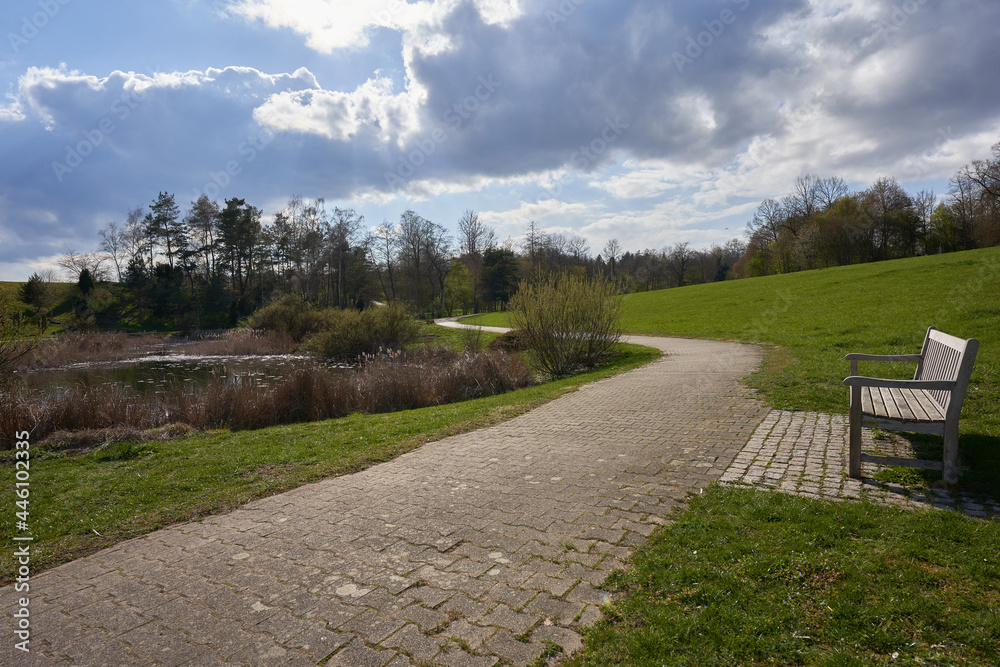 Path of gray cobblestone leads around a lake. Park bench nearby. Lots of green grass and various shrubs. Blue sky with clouds.