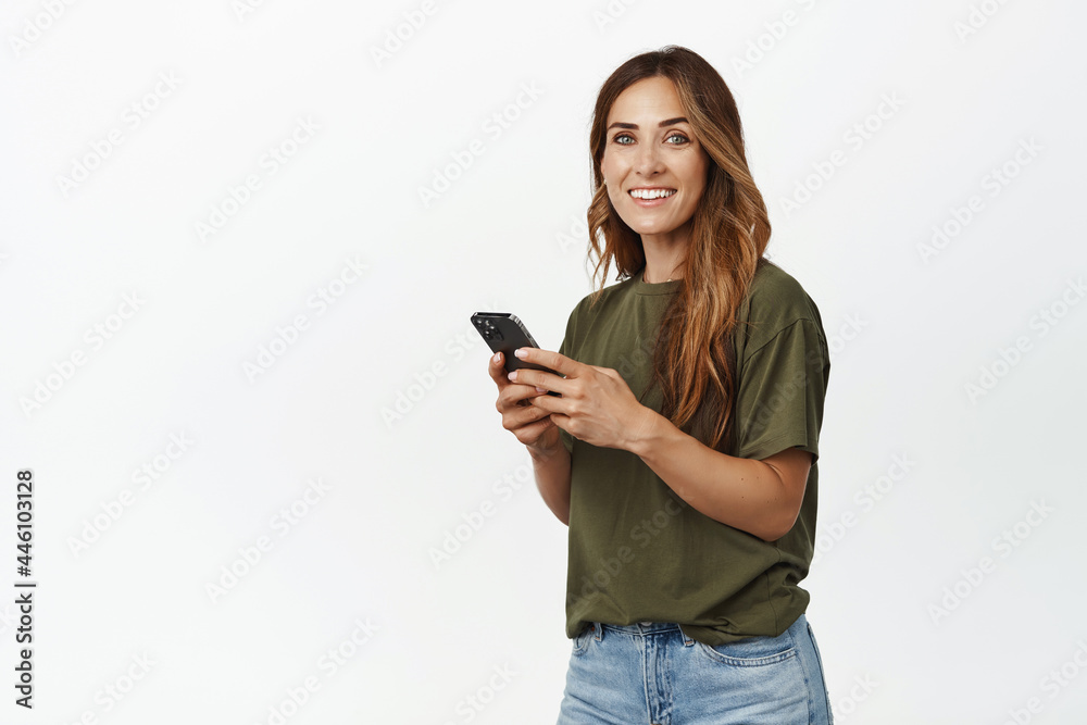Smiling adult woman using cellular, chat on mobile phone, holding smartphone, standing in tshirt and jeans against white background