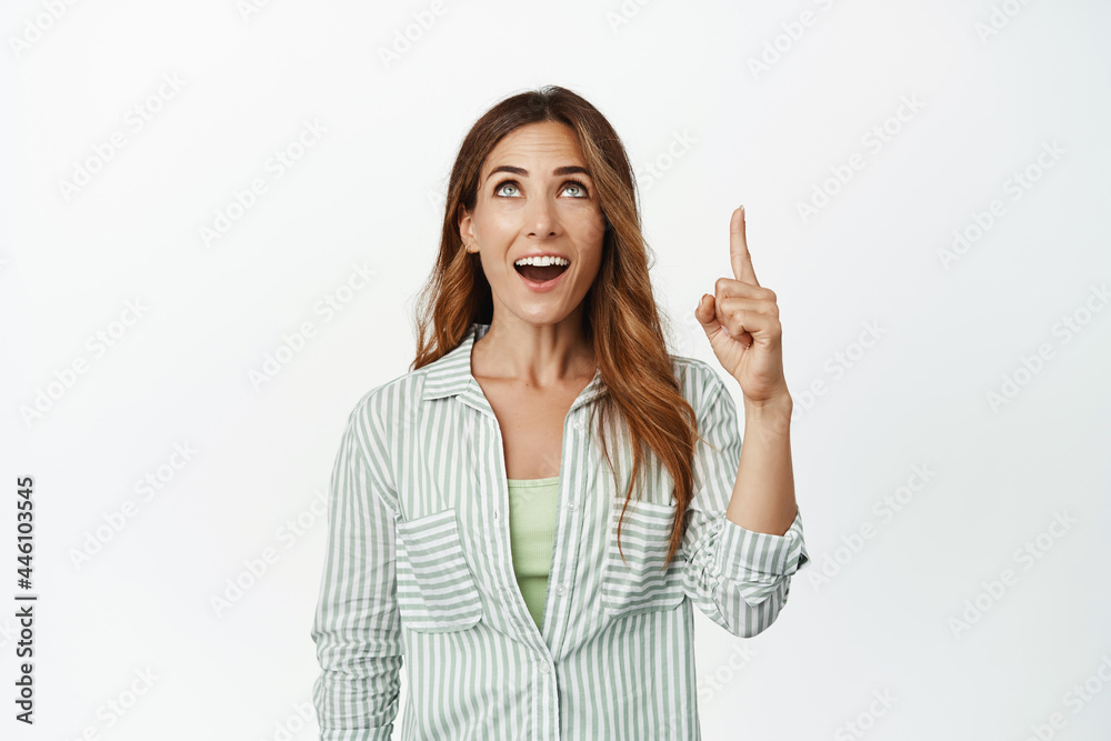 Image of excited, amazed brunette woman, gasp with WOW face, pointing finger and looking up at smth awesome, like promo deal, standing against white background Image of excited, amazed brunette woman, gasp with WOW face, pointing finger and looking up at smth awesome, like promo deal, standing against white background