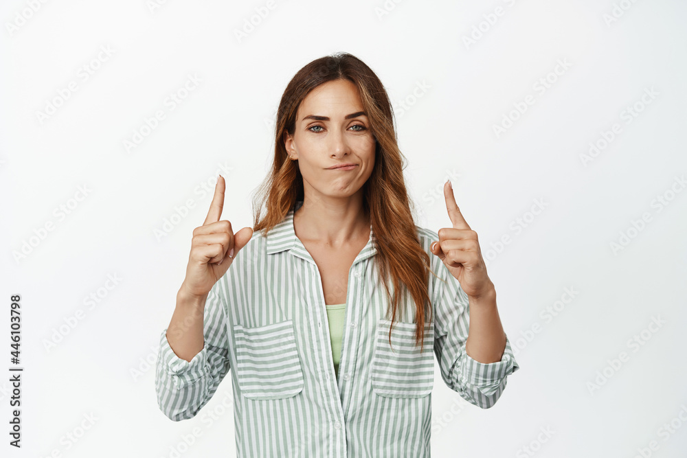 Smug face. Confident and ambitious business woman smiling pleased, pointing fingers up at top promo sale, showing advertisement proud, standing over white background