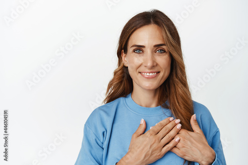 Portrait of smiling brunette woman keep hands on heart and looking caring, grateful, gratitude expression, standing in blue blouse against white background © Cookie Studio