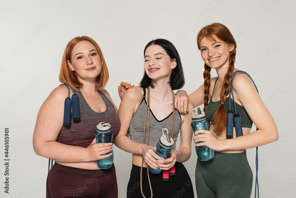 Body positive sportswomen with sports bottles and jump ropes smiling isolated on grey Body positive sportswomen with sports bottles and jump ropes smiling isolated on grey