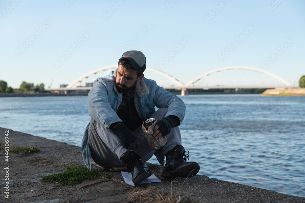 Young depressed suicidal drunk homeless man sitting under the bridge drinking alcohol drink and thinking to jump in to the river to commit suicide and end his life, social documentary concept