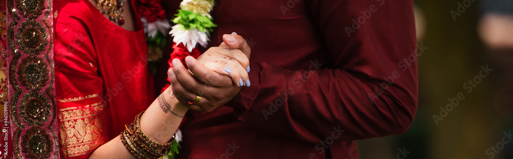 cropped view of indian man holding hands with bride with mehndi in sari, banner