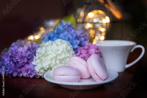 Pink marshmallow on a white saucer with multicolored hydrangea flowers and a cup against the background of evening lights. Summer evening, holiday mood concept.