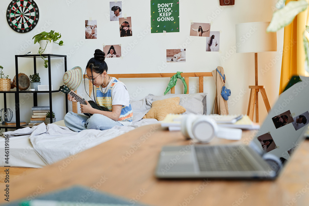 Obraz premium Portrait of Asian teenage girl playing ukulele while sitting on bed in cozy room, copy space