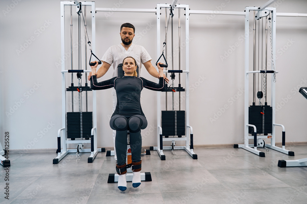 Fototapeta premium Kinesiologist helps young woman doing exercises to strengthen back muscles. Female patient treatment