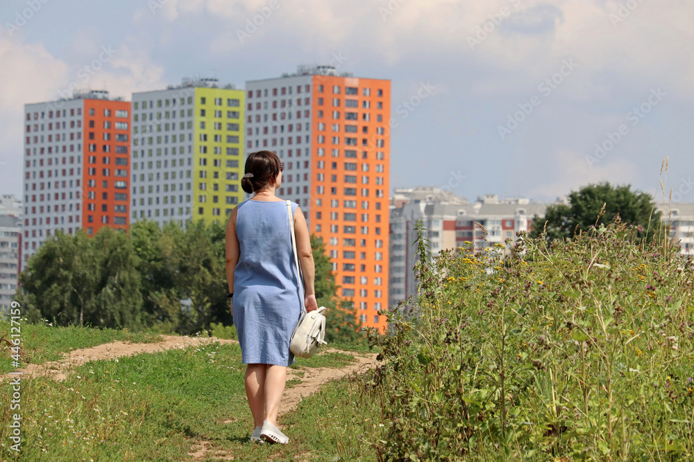 Woman walking on green field and looking to new residential buildings. Real estate, choosing home or renting property at summer