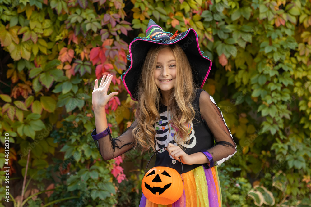 Hello Halloween holiday. A girl in a witch costume holds a basket of pumpkin jack and waves joyfully, standing against the background of autumn trees.