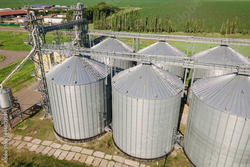 Metal silos on field aerial view. Large containers for storing and processing grains. Silver grain elevators in farmland. Storage tank view from above.