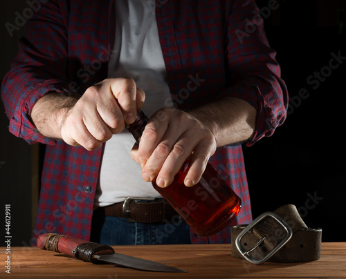 A man in a rustic-looking check shirt tries to open a bottle of whiskey. alcoholism and aggression concept. close-up.