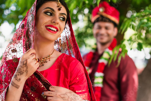 happy indian bride in sari and headscarf near blurred man in turban on background