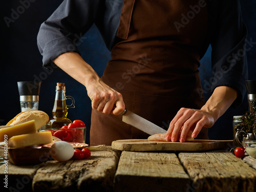 The process of slicing chicken fillet for making Caesar salad. A rough wooden table on which the ingredients for the salad are laid out. Delicious restaurant dish. Home kitchen.