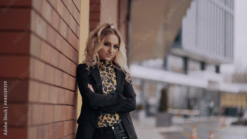 Leaning against the wall of a building, the sexy and serious blonde joins her hands to her chest, has a strident make-up, dressed in office. Female energy. Beauty industry. Artistic concept Leaning against the wall of a building, the sexy and serious blonde joins her hands to her chest, has a strident make-up, dressed in office. Female energy. Beauty industry. Artistic concept