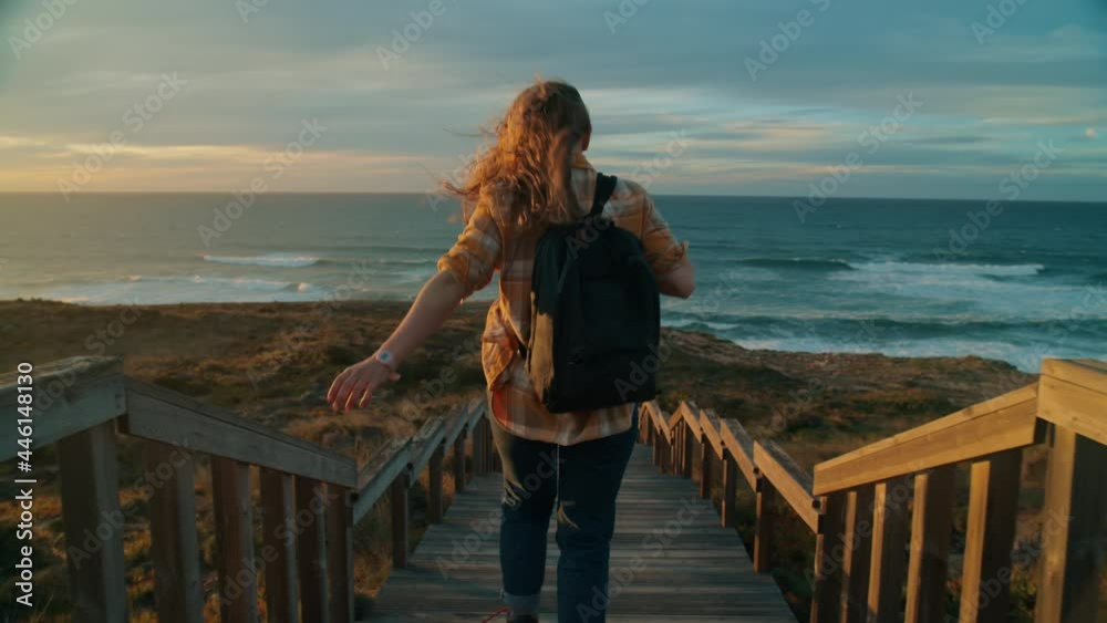 Cinematic inspiring shot of young woman run down epic boardwalk down to rocky ocean coastline. Female adventurer explore sunset coast. Scenic vacation travel destination. Wanderlust lifestyle