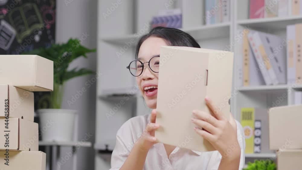 Happy young Asian woman wearing glasses and sitting to do live on social media at home, holding and present her product by camera, Women doing business selling online, woking at home Happy young Asian woman wearing glasses and sitting to do live on social media at home, holding and present her product by camera, Women doing business selling online, woking at home