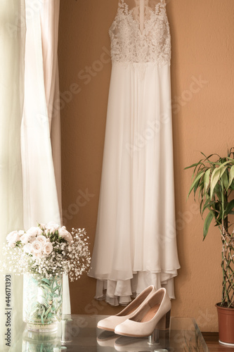 vertical shot of a beautiful wedding slippers on a glass table and a floral arrangement with white flowers and a wedding dress in the background on the wall