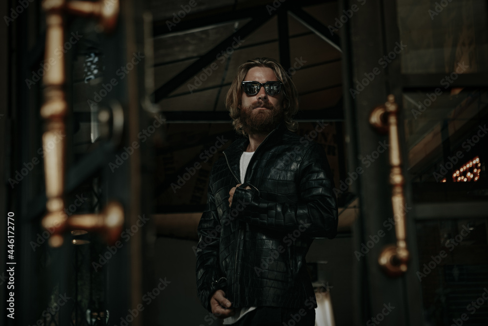 Brutal caucasian  man with long beard hipster in black leather jacket stay next to glass door 