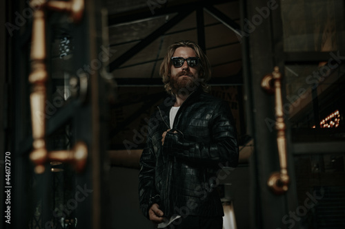 Brutal caucasian man with long beard hipster in black leather jacket stay next to glass door 