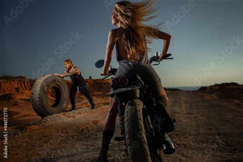 Fototapeta Naklejka Na Ścianę i Meble -  Sexy young women sit on motorcycle and looking at man rolling the tire at sunset.