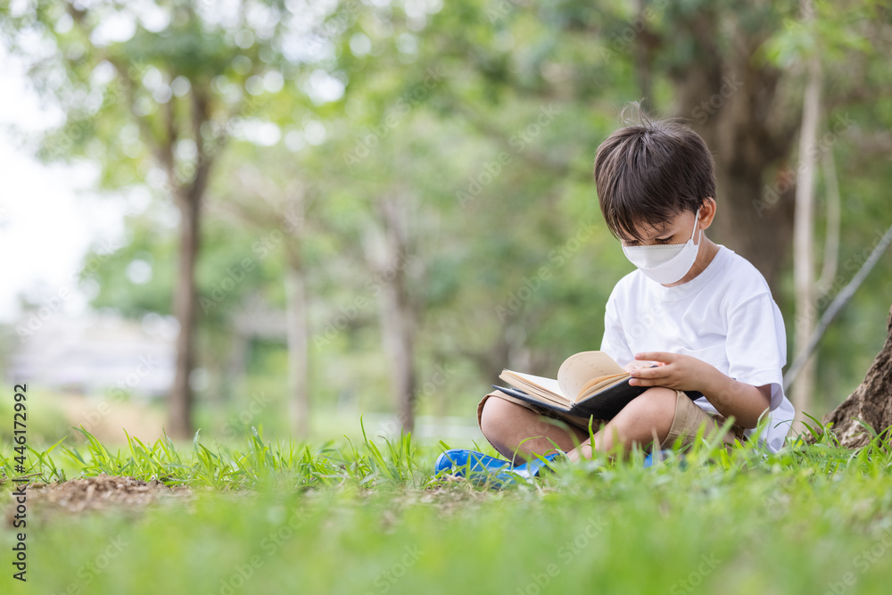 A white boy wearing a hygienic mask sits alone under a tree reading a book.