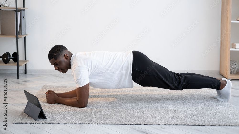 Online training. Sportive black man. Strong body. Quarantine lifestyle. Happy african guy doing plank workout looking portable computer light room interior. Online training. Sportive black man. Strong body. Quarantine lifestyle. Happy african guy doing plank workout looking portable computer light room interior.