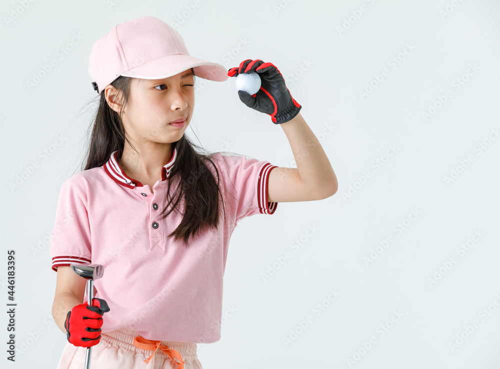 Close up isolated studio shot of Asian little professional girl kid golfer in pink athlete fashion clothing red gloves and cap stand hold driver and golf ball close eye aiming for target on green