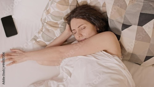 Top view of dark-haired Caucasian woman sleeping in bedroom, trying groping with hand and eyes closed her cellphone on bed, then frowning and getting back to sleep photo