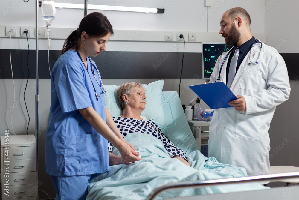 Nurse reading blood oxygen saturation from oxymeter device attached on sick senior woman patient laying in hospital bed resting. Doctor monitoring patient health condition. Nurse reading blood oxygen saturation from oxymeter device attached on sick senior woman patient laying in hospital bed resting. Doctor monitoring patient health condition.
