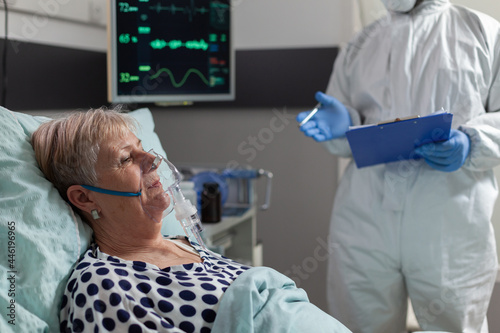 Sick senior woman inhale and exhale through oxygen mask laying in hospital bed during global pandemic with coronavirus. Doctor dressed in coverall pee suit to prevent infection with covid-19.