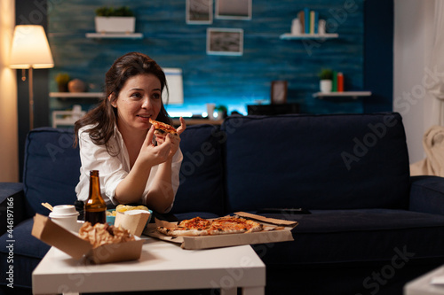 Caucasian female holding delicious pizza slice eating takeaway food delivery while watching comedy film on television at night. Woman enjoying junk-food home delivered relaxing on couch