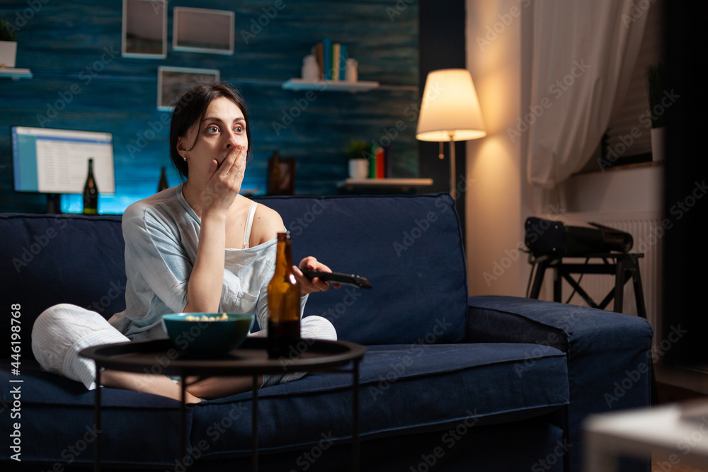 Focused interested woman eating popcorn watching interesting movie show on television. Female sitting on comfortable couch dressed in pajama late at night in living room having concentrated expression Focused interested woman eating popcorn watching interesting movie show on television. Female sitting on comfortable couch dressed in pajama late at night in living room having concentrated expression