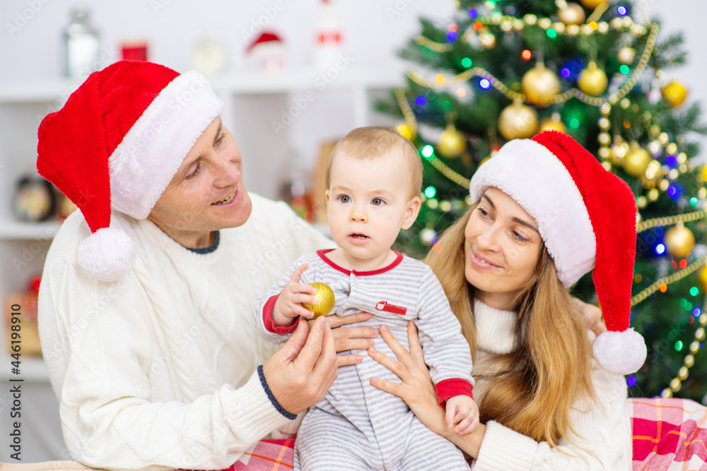Young couple in santa hats hold their son in their arms against the background of a christmas tree at home Young couple in santa hats hold their son in their arms against the background of a christmas tree at home