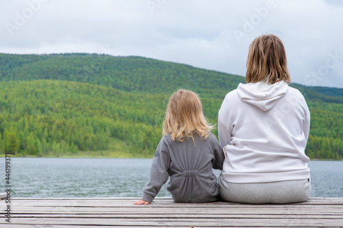Mom hugs the child sitting next to her on the pier of a blue lake in the mountains and gently looks at the child. Family tourism