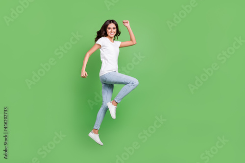 Full size profile photo of cool brunette young lady jump hands fists wear t-shirt jeans isolated on green color background