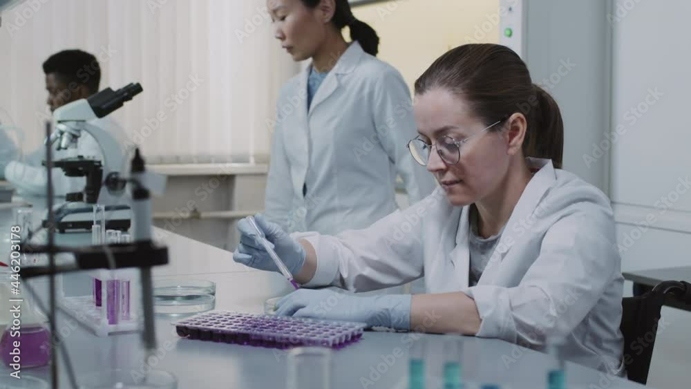 Slowmo tracking portrait of disabled female chemist in wheelchair putting solution into petri dish and posing for camera Scientists in white coats doing research in background