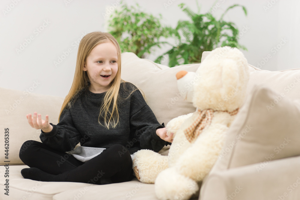 Photo of teddy bear and blonde girl talking with each other on the sofa.