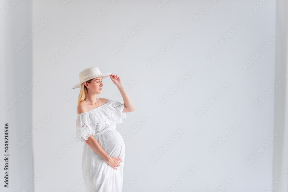 Young pregnant woman in anticipation of a baby in a white hat, dress, hugs her belly. Tender blonde girl in a minimalistic, isolated interior of airy fabrics. Happy maternity pregnancy, surrogacy Young pregnant woman in anticipation of a baby in a white hat, dress, hugs her belly. Tender blonde girl in a minimalistic, isolated interior of airy fabrics. Happy maternity pregnancy, surrogacy