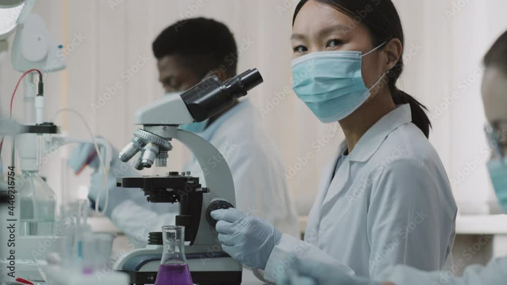 Tilt up slowmo shot of Asian female scientist in face mask, gloves and white coat using microscope, then turning towards camera while working in lab with her colleagues Tilt up slowmo shot of Asian female scientist in face mask, gloves and white coat using microscope, then turning towards camera while working in lab with her colleagues