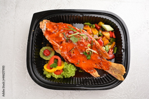 deep fried crispy whole grouper fish seafood with sweet and sour chilli sauce and lychee fruits on top in black bento tray box for asian take away halal menu