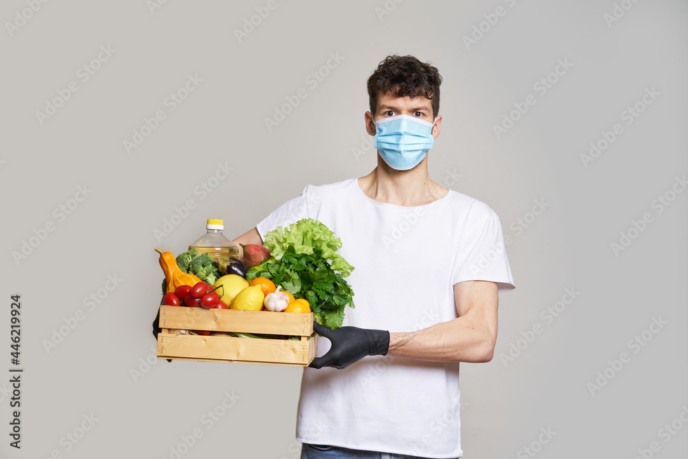 Food delivery concept during quarantine. A man in a mask, gloves and a white T-shirt holds a box of fresh food in his hands. Food delivery from the supermarket, from farmers home. Food delivery concept during quarantine. A man in a mask, gloves and a white T-shirt holds a box of fresh food in his hands. Food delivery from the supermarket, from farmers home.