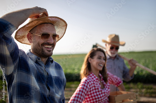 Three farmers walking in field carrying crate with produce and hoe smiling. © Budimir Jevtic