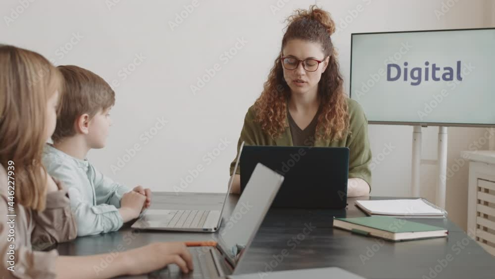 Tracking left medium close-up of young Caucasian woman wearing glasses, teaching elementary-aged boy and girl sitting at table, using computers on lesson, smiling Tracking left medium close-up of young Caucasian woman wearing glasses, teaching elementary-aged boy and girl sitting at table, using computers on lesson, smiling