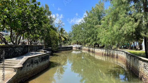 Winding river and a beautifully laid out stone embankment in summer. City park, white bridge over the river. No people. Green trees along walking road. Reflection of green spaces on a water surface.