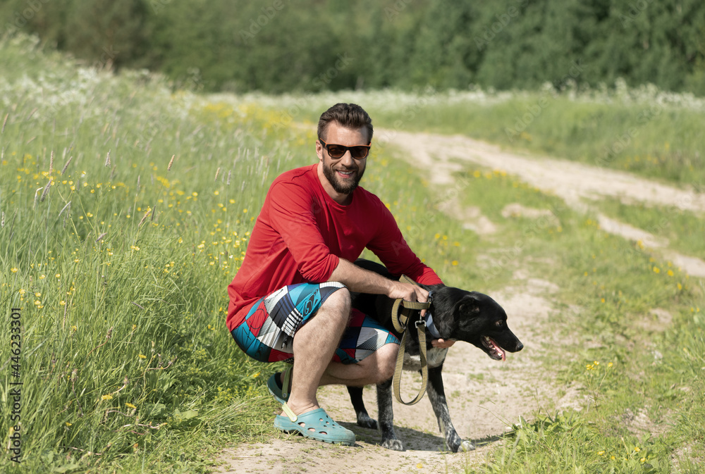 Caucasian smiling man, 30 years old, and his black dog are on a rural road. Caucasian smiling man, 30 years old, and his black dog are on a rural road.