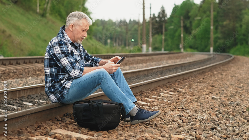An elderly male tourist is sitting on the railway tracks, looking through information on the phone and waiting for the train. A middle-aged tourist waits for a train on the railway on a summer day. An elderly male tourist is sitting on the railway tracks, looking through information on the phone and waiting for the train. A middle-aged tourist waits for a train on the railway on a summer day.