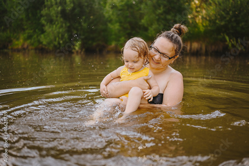 Little funny cute blonde girl child toddler in yellow bodysuit laughing learns swim outside at summer lake. Plus size body positive smiling woman mother baby swimming in natural pool. Water splashes