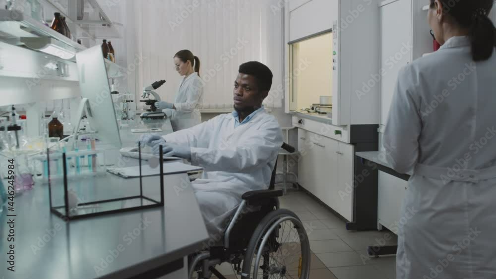 Slowmo tracking of disabled black male scientist in wheelchair working on computer in lab, then posing His female colleagues doing their research in background White lab rat is in glass tank Slowmo tracking of disabled black male scientist in wheelchair working on computer in lab, then posing His female colleagues doing their research in background White lab rat is in glass tank