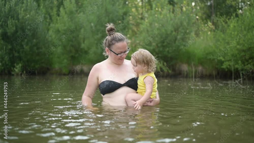 Little funny cute blonde girl child toddler in yellow bodysuit laughing learns swim outside at summer lake. Plus size body positive smiling woman mother baby swimming in natural pool. Water splashes Little funny cute blonde girl child toddler in yellow bodysuit laughing learns swim outside at summer lake. Plus size body positive smiling woman mother baby swimming in natural pool. Water splashes