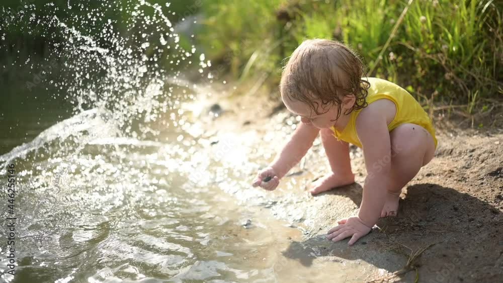 Little funny cute blonde girl child toddler in yellow wet bodysuit playing by the lake waterside shore at sunset outside. Baby with mud on hands and legs. Water activities at summer Little funny cute blonde girl child toddler in yellow wet bodysuit playing by the lake waterside shore at sunset outside. Baby with mud on hands and legs. Water activities at summer